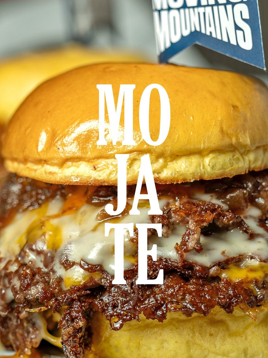 Primerísimo plano de una hamburguesa de Dughi's: pan brillante, queso fundido y el borde de la carne caramelizado. Sobre la imagen, la palabra MÓJATE escrita en vertical.